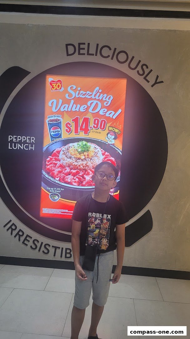 A young boy stands in front of a Pepper Lunch advertisement featuring a sizzling value deal.