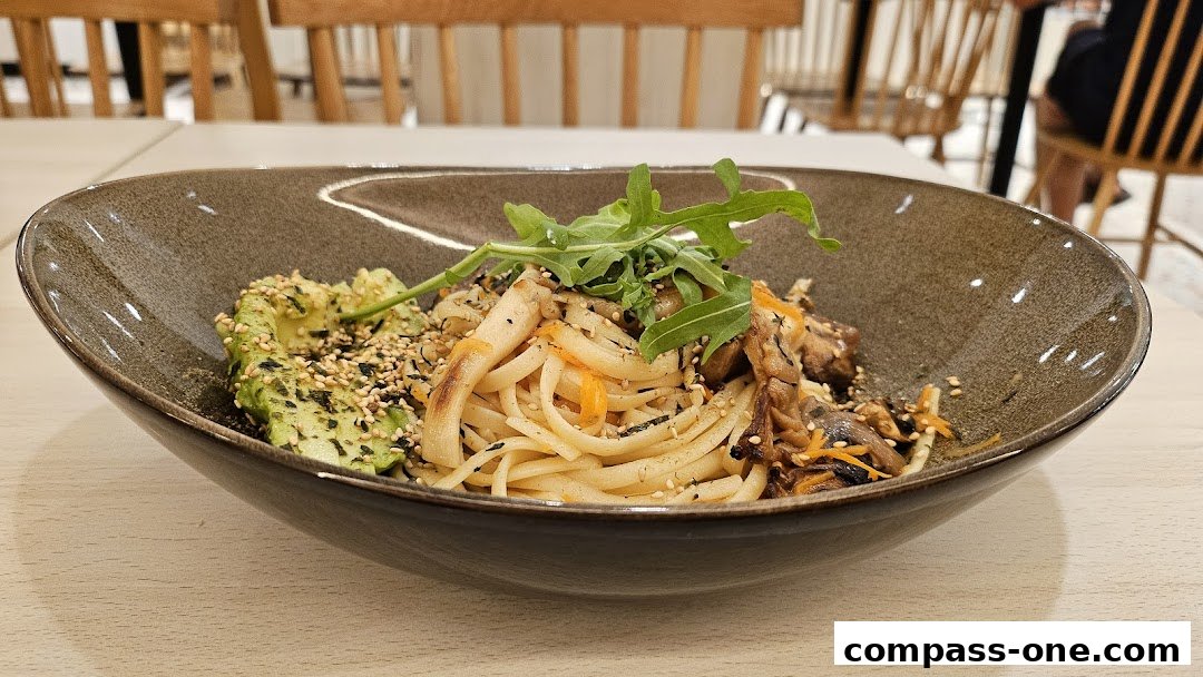 A bowl of noodles with avocado, mushrooms, and greens, garnished with sesame seeds.