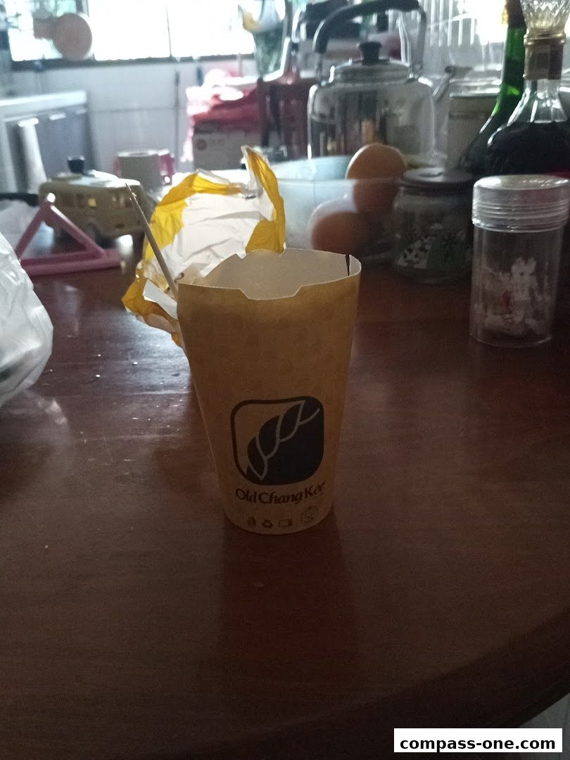 A paper cup with the Old Chang Kee logo sits on a wooden table with various items in the background.