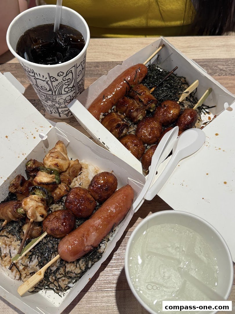 Two takeaway boxes filled with rice, skewers of meat, and sausages, alongside drinks.