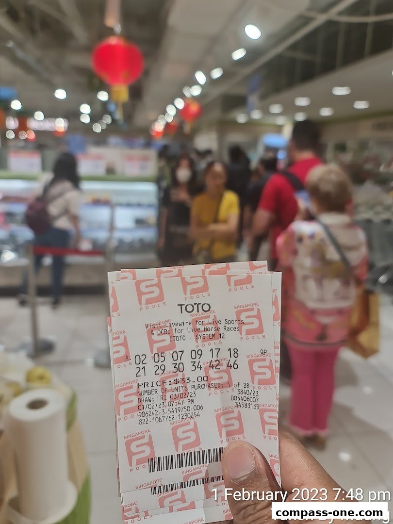 A hand holds Singapore Pools TOTO lottery tickets inside a brightly lit store.