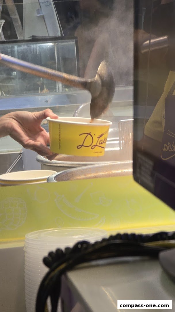 A hand holds a yellow cup with the D'Laksa logo while a ladle serves food.