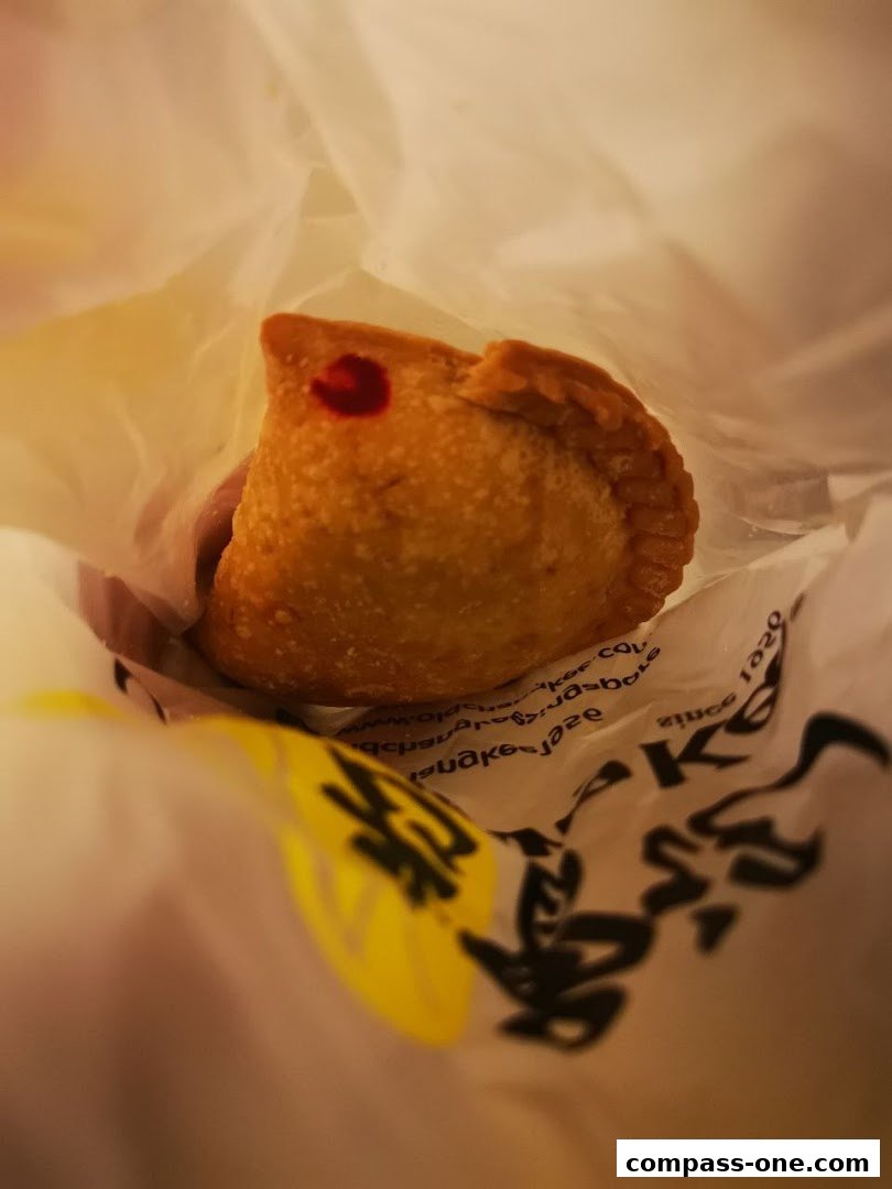 A close-up shot of a golden-brown curry puff with a red dot, held in a white paper bag.