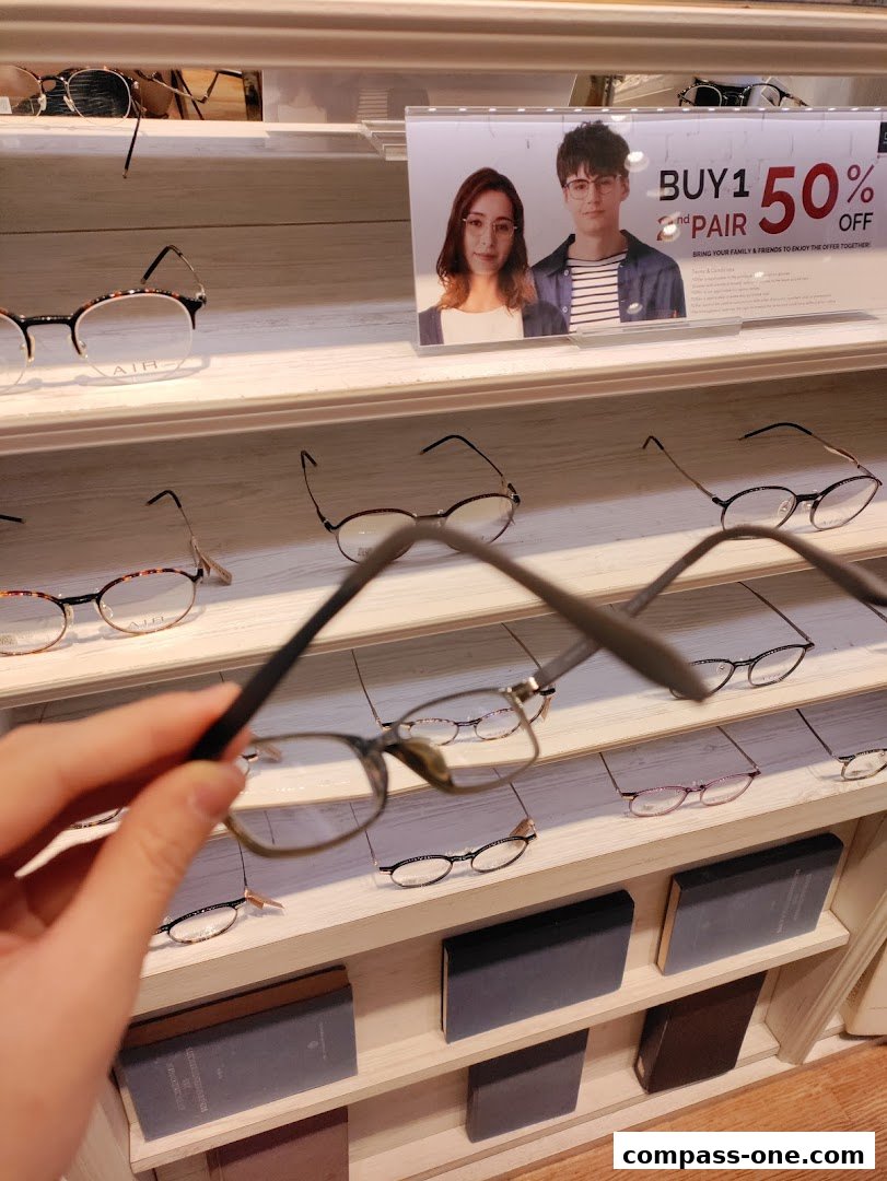 A hand holds up a pair of glasses in front of a display of eyeglasses and a promotional sign.