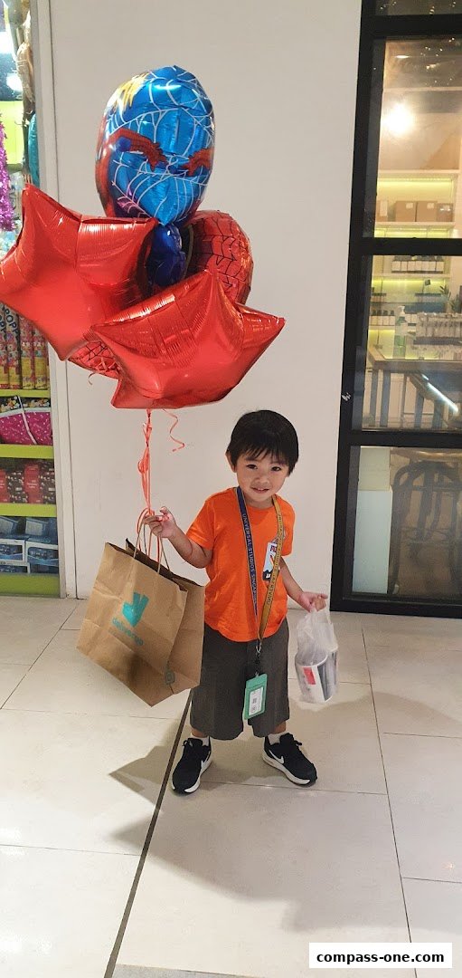 A young boy in an orange shirt holds Spiderman balloons and shopping bags.