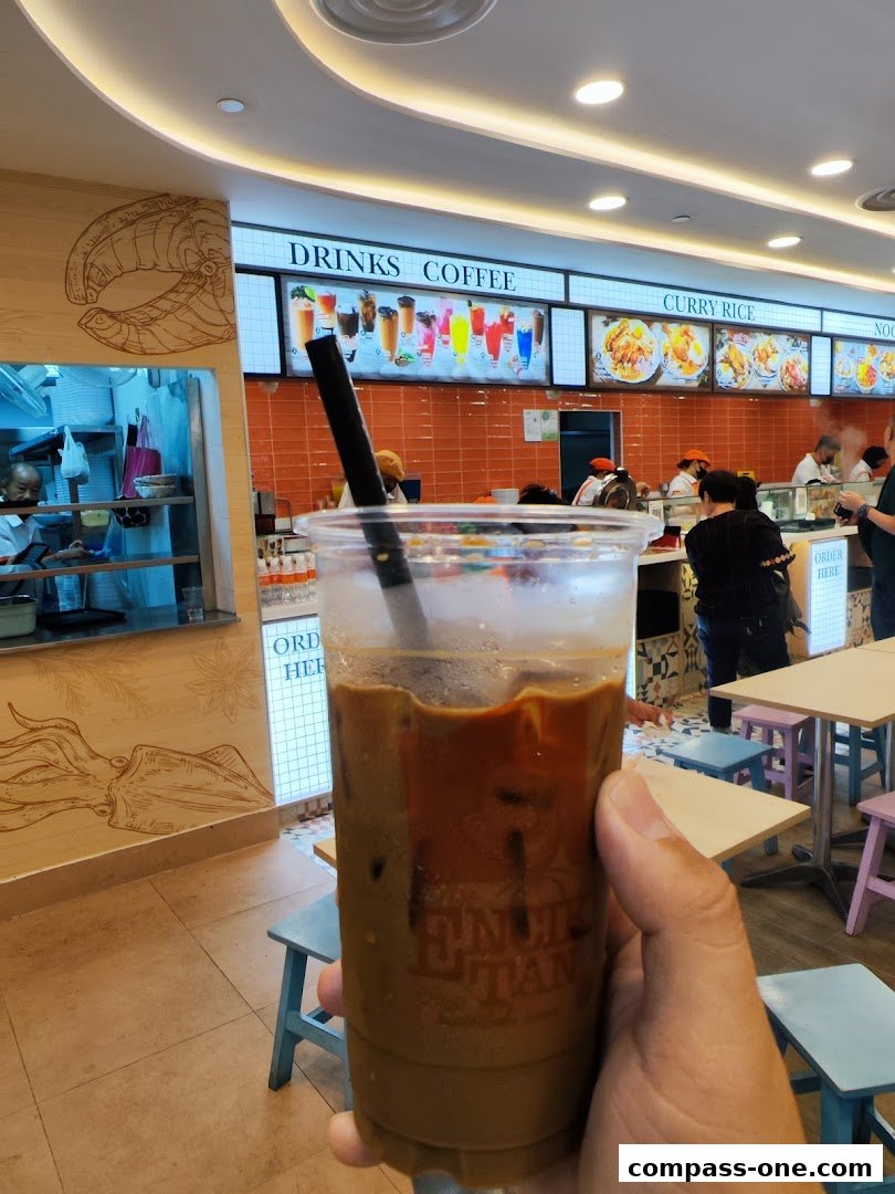 A person holds a coffee drink in front of a brightly lit food stall with menu displays.