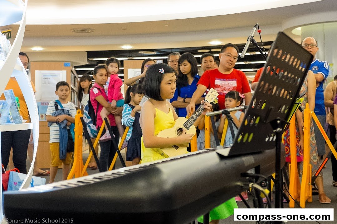 A young girl plays the ukulele at a music school event with an audience watching.