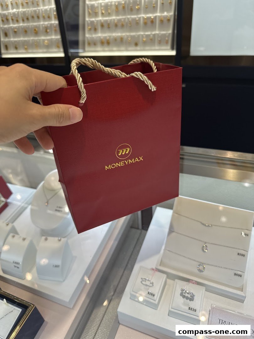 A hand holds a red MoneyMax shopping bag in front of a jewelry display.