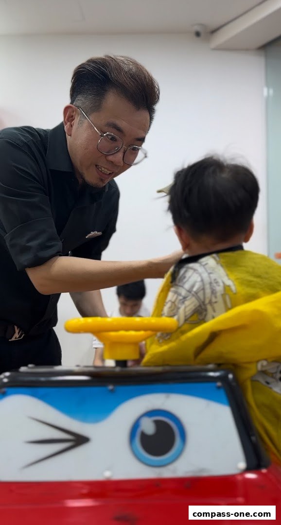A barber cuts a child's hair while the child sits in a toy car.