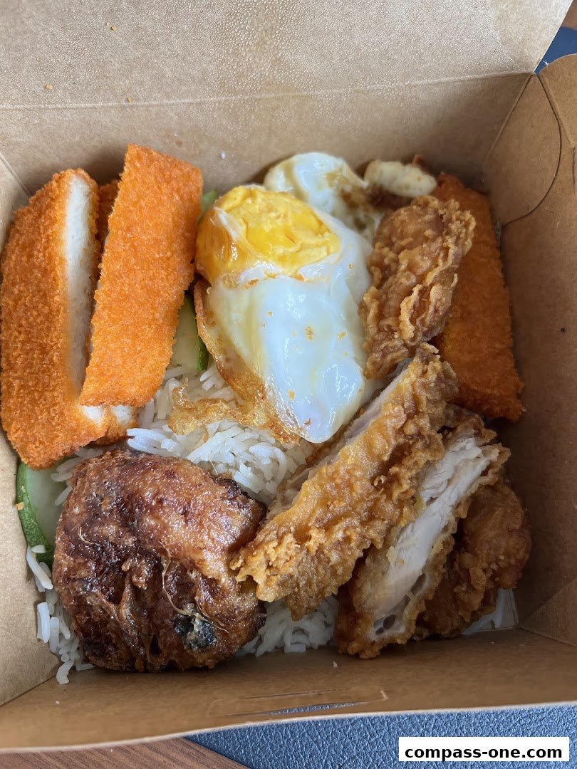 A close-up of a takeaway box filled with nasi lemak, featuring fried chicken, egg, and fish.
