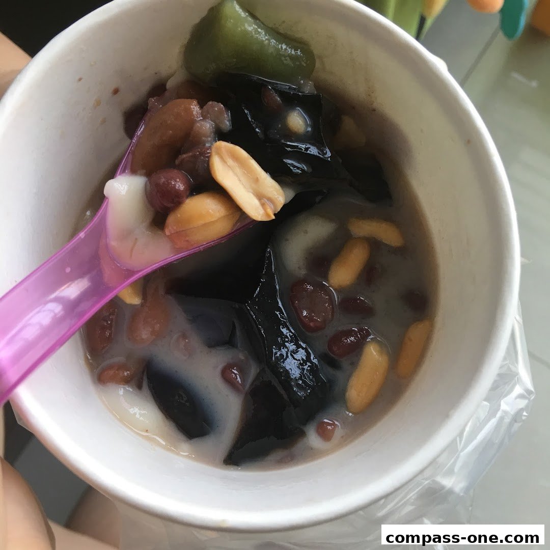A close-up of a white cup filled with a refreshing dessert, featuring grass jelly, red beans, peanuts, and a green jelly.