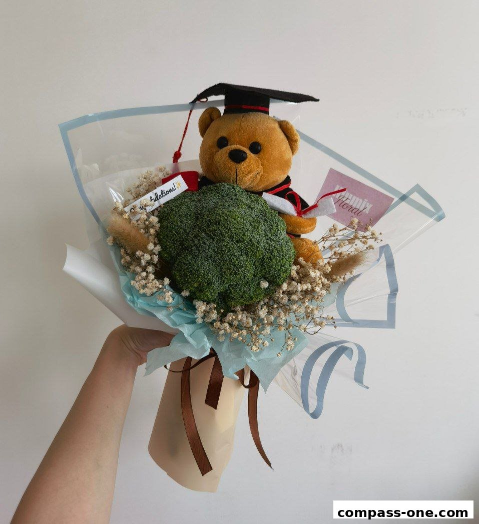 A hand holds a unique graduation bouquet featuring a teddy bear, broccoli, and baby's breath.