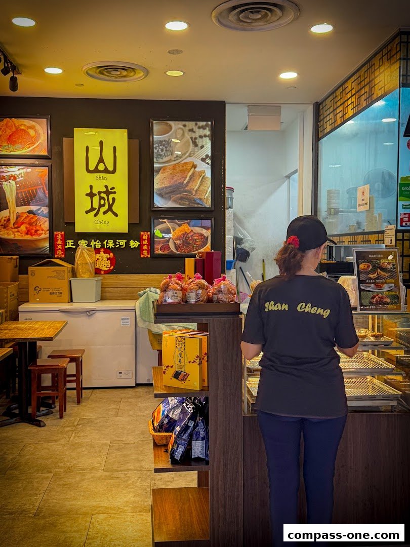 A person stands behind a counter in a shop selling Ipoh delicacies, with food images on the wall.