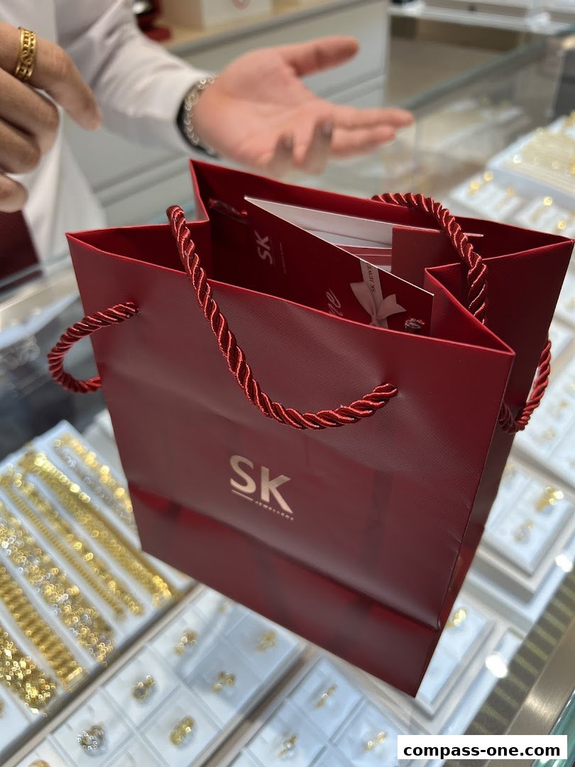 A red shopping bag with the SK Jewellery logo sits in front of display cases filled with jewelry.
