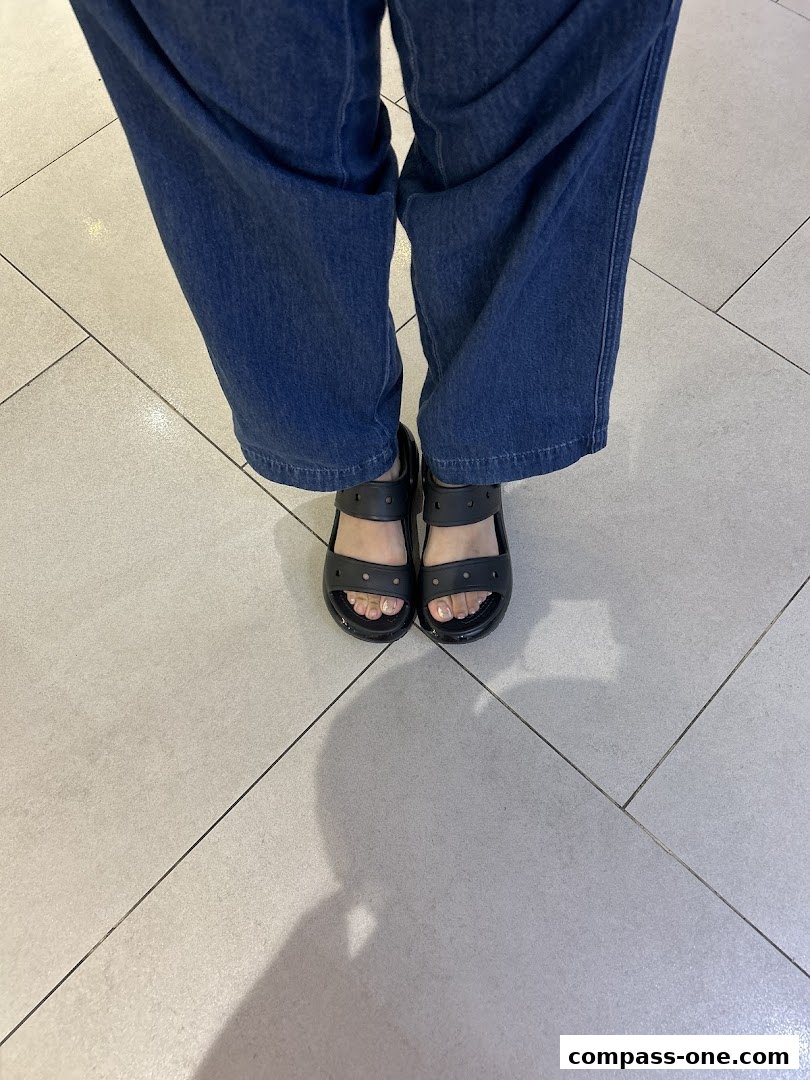A person wearing blue jeans and black Crocs sandals stands on a tiled floor.