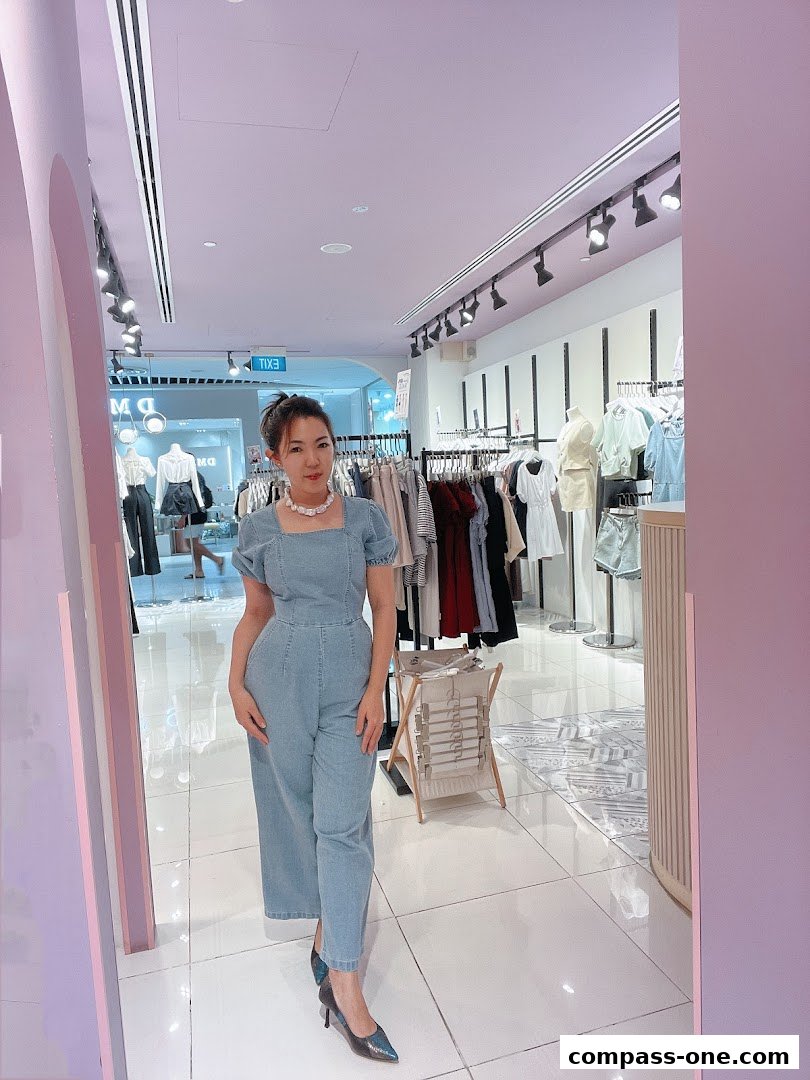 A woman models a denim jumpsuit inside a brightly lit clothing store.