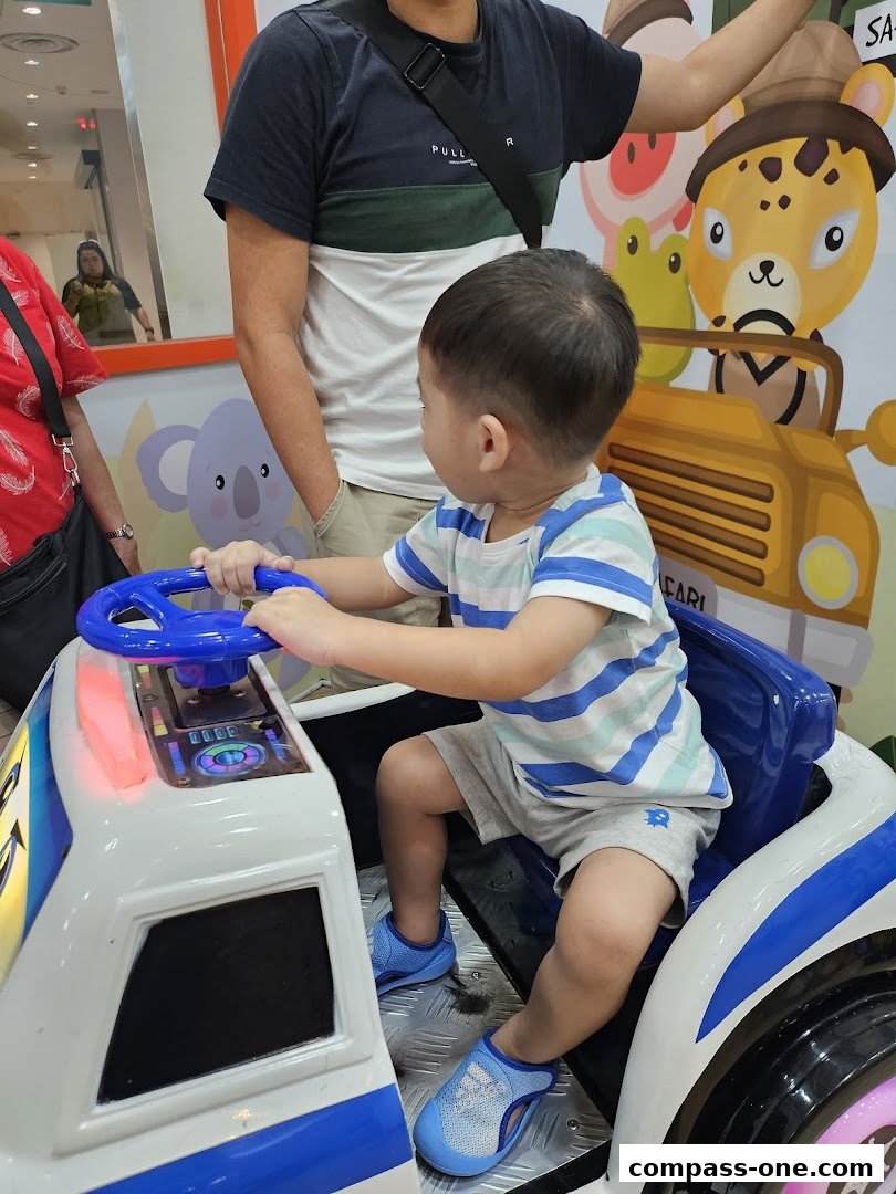 A young boy sits in a toy car, playing with the steering wheel, with adults nearby.