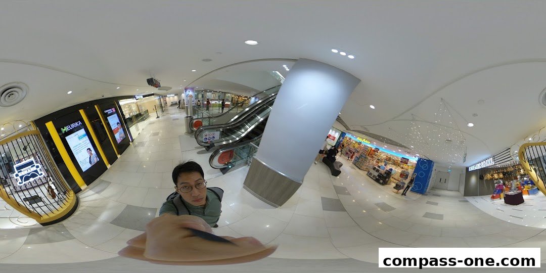 A person takes a selfie in a shopping mall hallway with escalators and shops.
