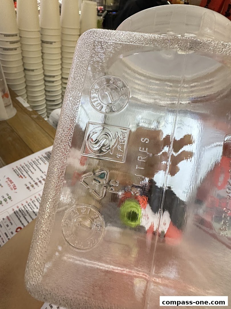 A close-up of a clear plastic container with recycling symbols and text, with a menu and stacked cups in the background.
