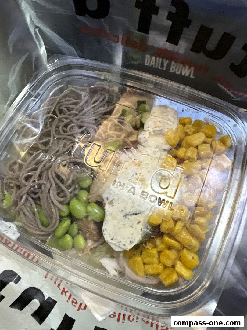 A clear plastic container filled with a healthy meal, featuring noodles, edamame, corn, and a creamy dressing.