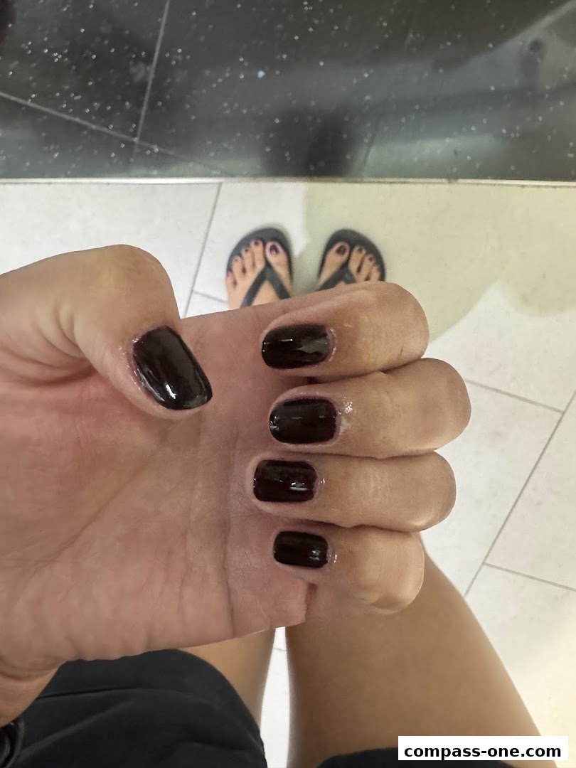 A close-up of a hand with dark, glossy nail polish and feet in flip-flops.