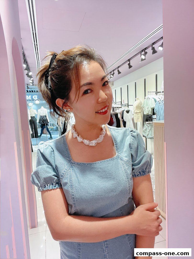 A woman smiles at the camera in a stylish denim dress and pearl necklace inside a boutique.