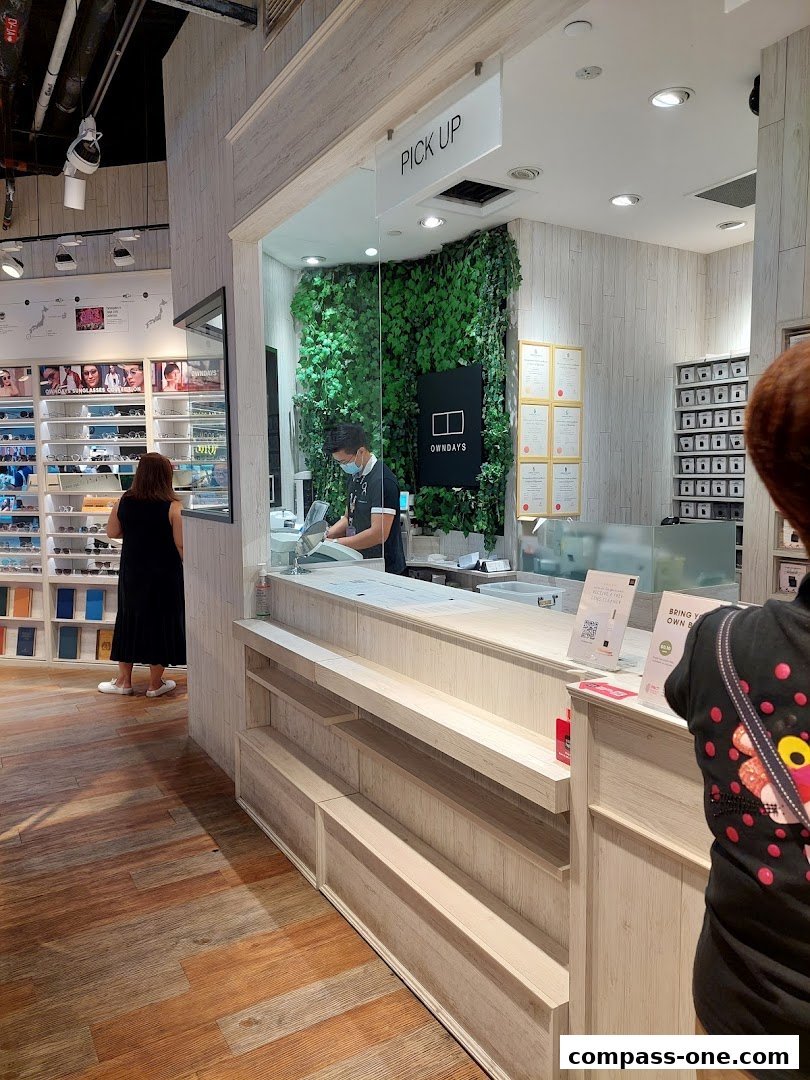 A customer picks up glasses at the OWNDAYS counter with a green wall and product displays.