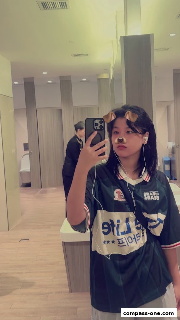 A person in a dark jersey with white lettering takes a mirror selfie in a clean, modern restroom.