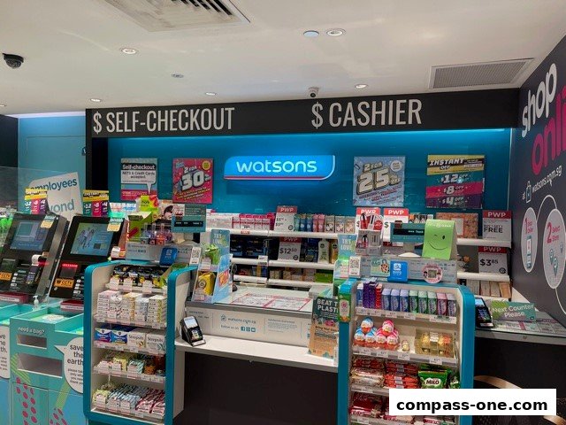 Interior view of a Watsons pharmacy with self-checkout and cashier counters, displaying various products and promotions.