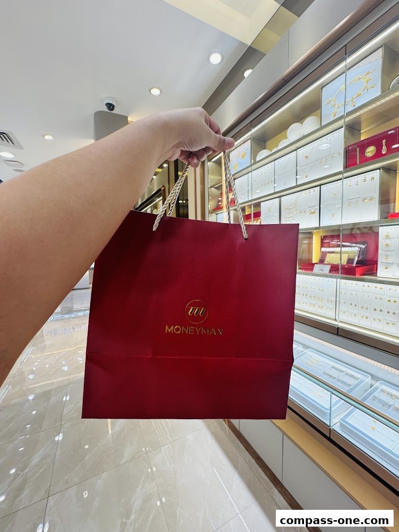 A hand holds a red shopping bag with the MoneyMax logo inside a jewelry store.