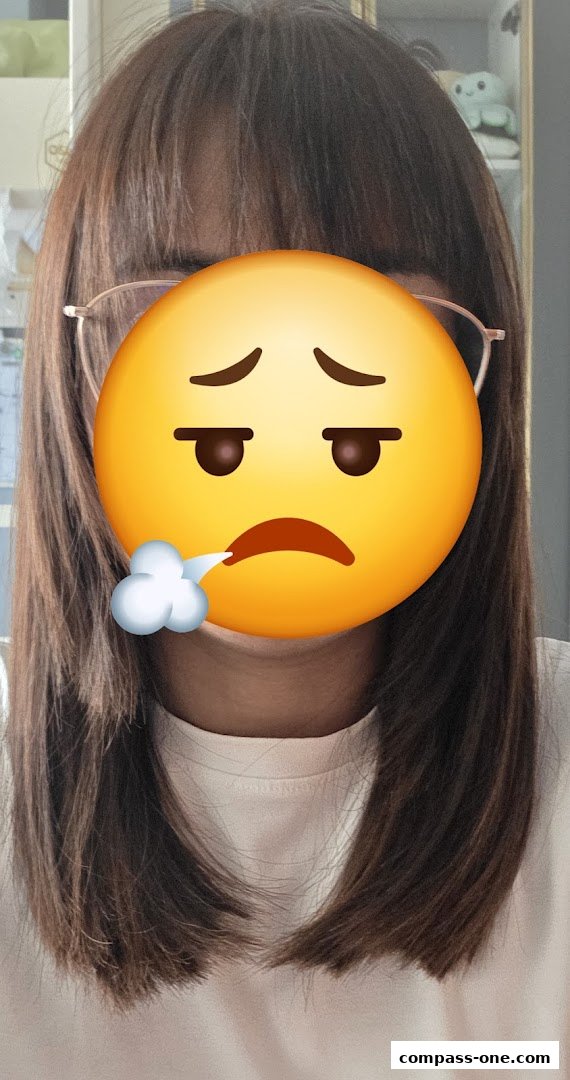 A person with brown hair and glasses, face covered by a sad emoji.