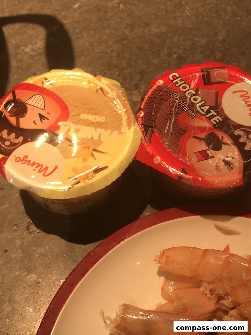 Two ice cream cups, one vanilla and one chocolate, sit on a surface next to a plate of shrimp.
