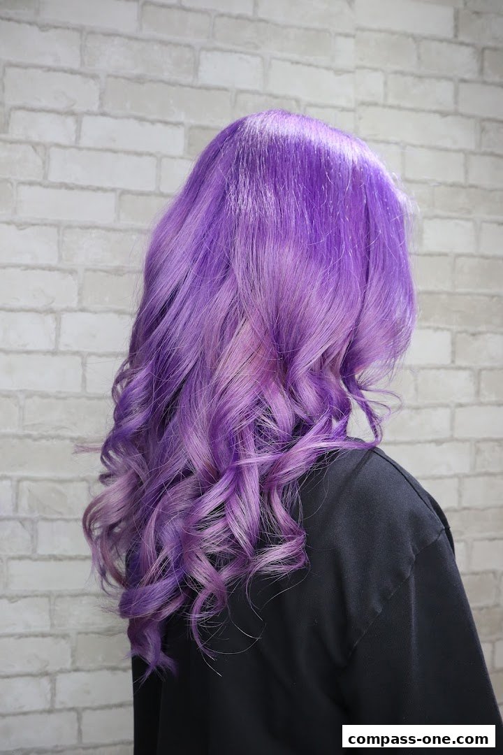 A person with vibrant purple, wavy hair styled against a brick wall.