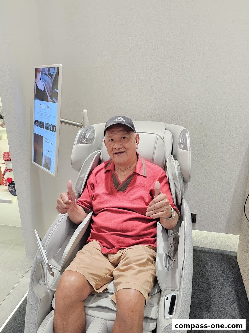 An elderly man sits in a massage chair, giving a thumbs up.