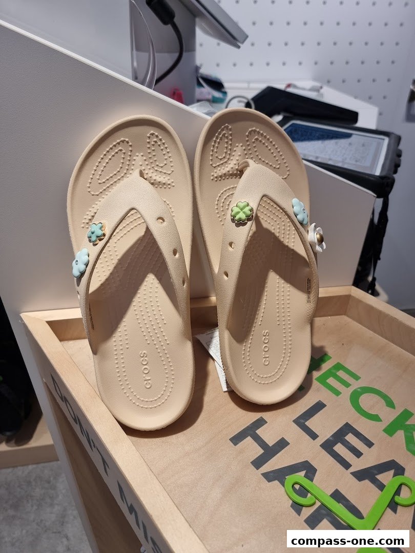 A pair of beige Crocs flip-flops with flower charms displayed on a wooden stand.