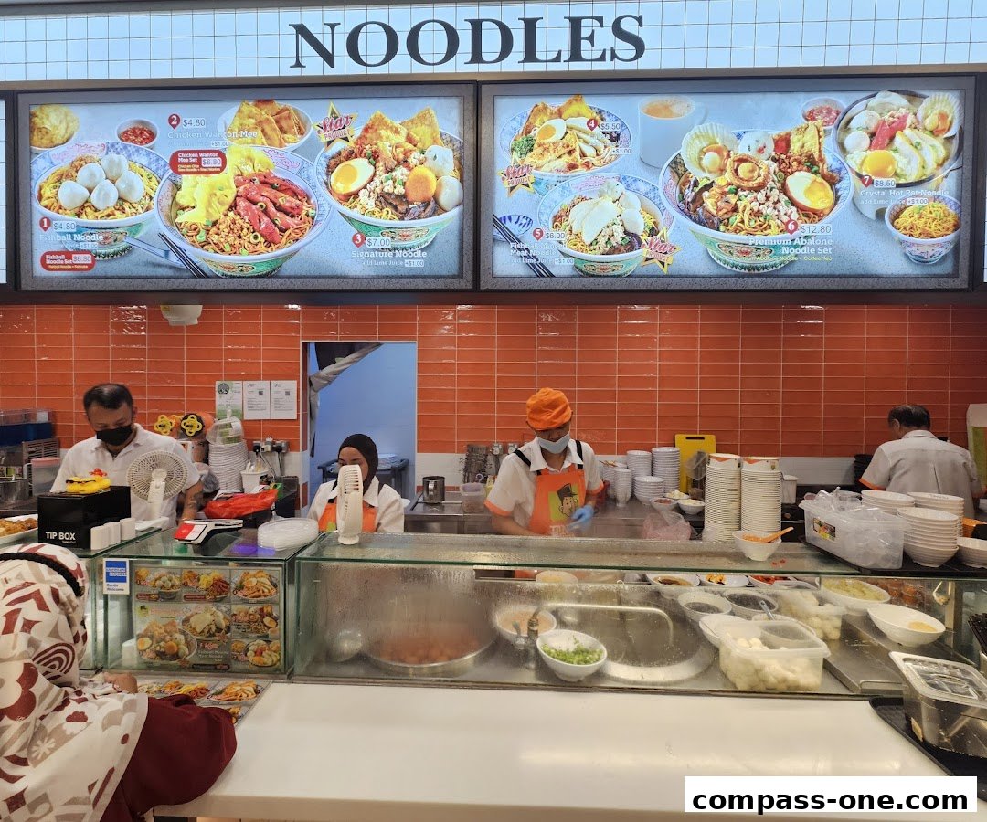 A food stall counter with a menu board displaying various noodle dishes and staff preparing orders.