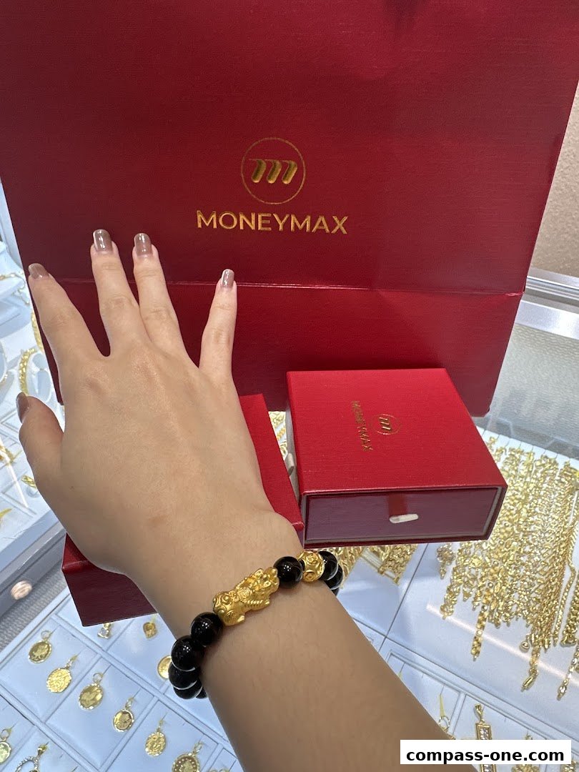 A hand wearing a gold and black beaded bracelet with a Pixiu charm in front of MoneyMax branded boxes.