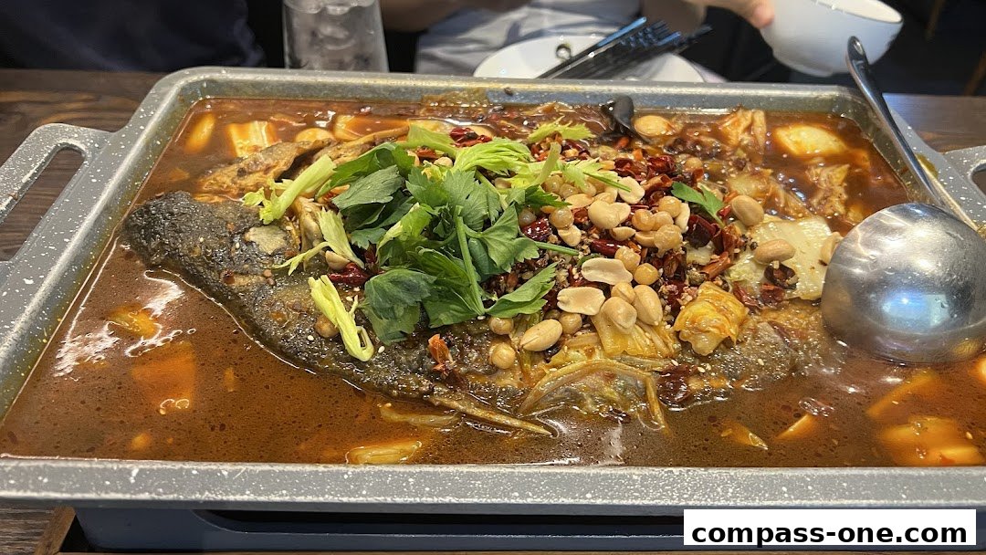 A close-up of a grilled fish dish served in a metal tray with broth, peanuts, and chili.