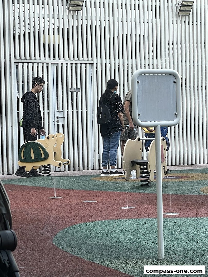 People stand near playground equipment with water spraying from the ground.