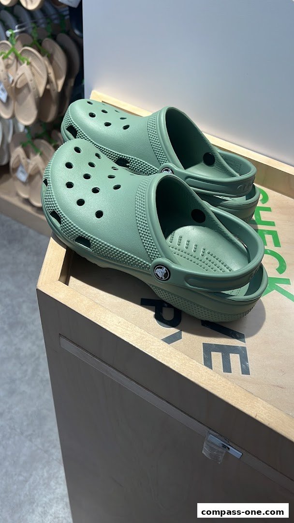 A pair of sage green Crocs clogs are displayed on a wooden stand.