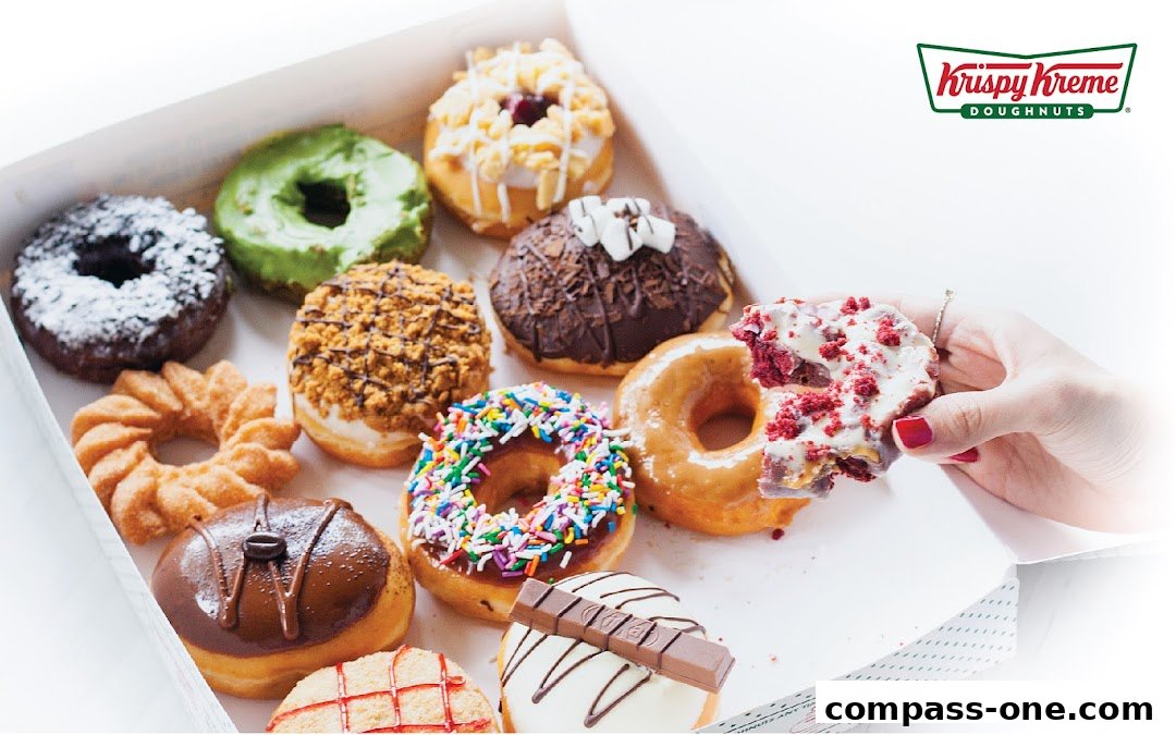 A box of assorted Krispy Kreme donuts, with a hand holding a red velvet donut.