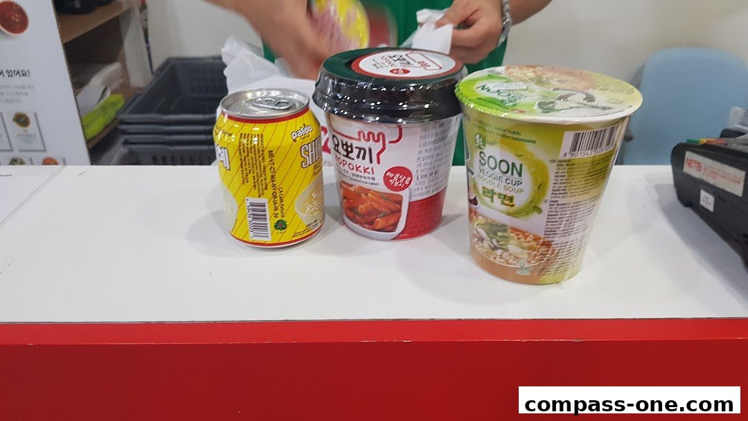 A selection of Korean instant food products, including a can of drink and two cup noodles, are displayed on a counter.