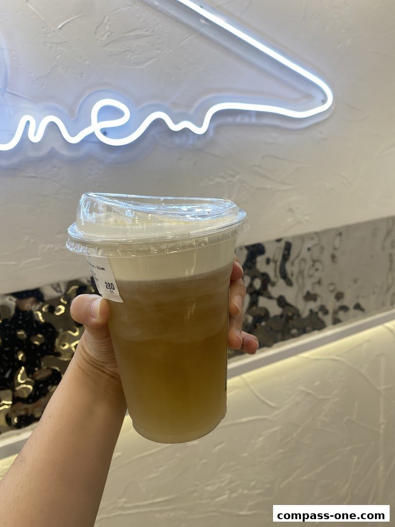A hand holds a clear plastic cup with a creamy topping and a light brown liquid.