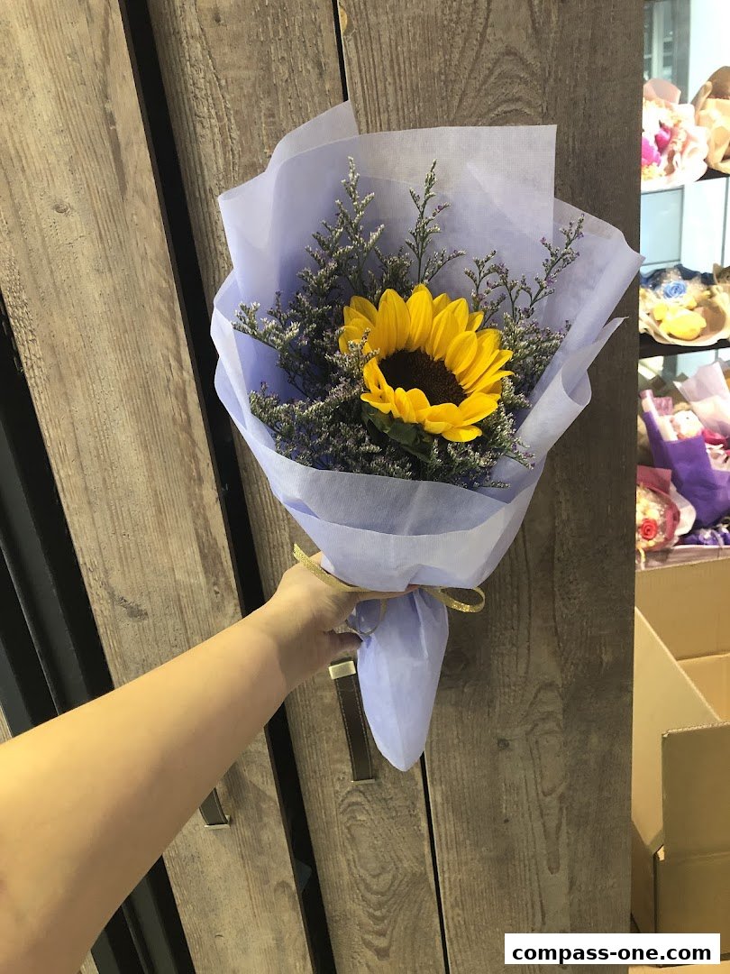 A hand holds a sunflower bouquet wrapped in light purple paper with a golden ribbon.