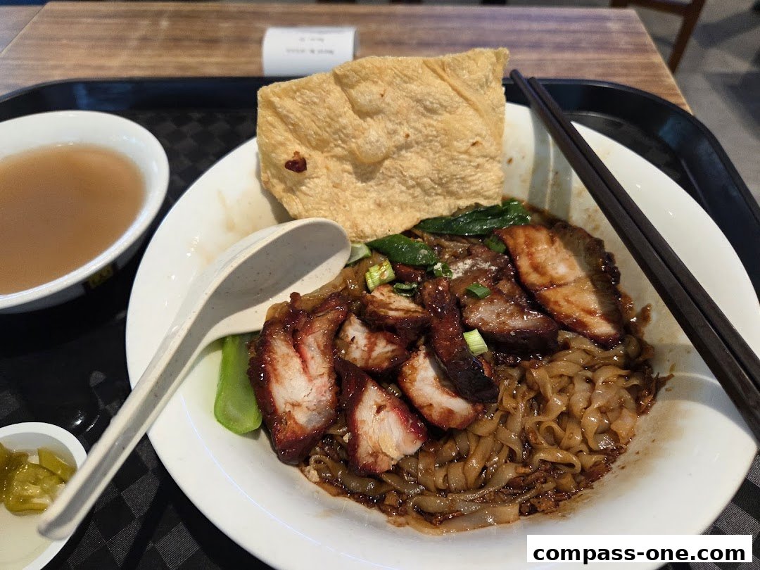 A bowl of Ipoh style noodles with char siu, greens, and a side of soup.