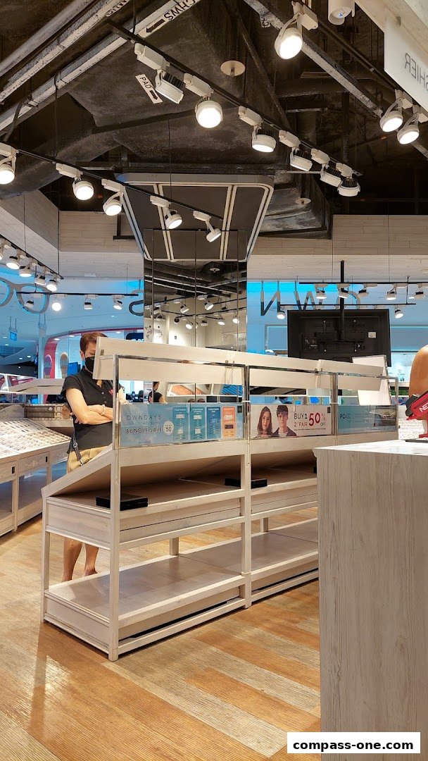 A woman stands in an optical shop with display shelves and promotional signs.