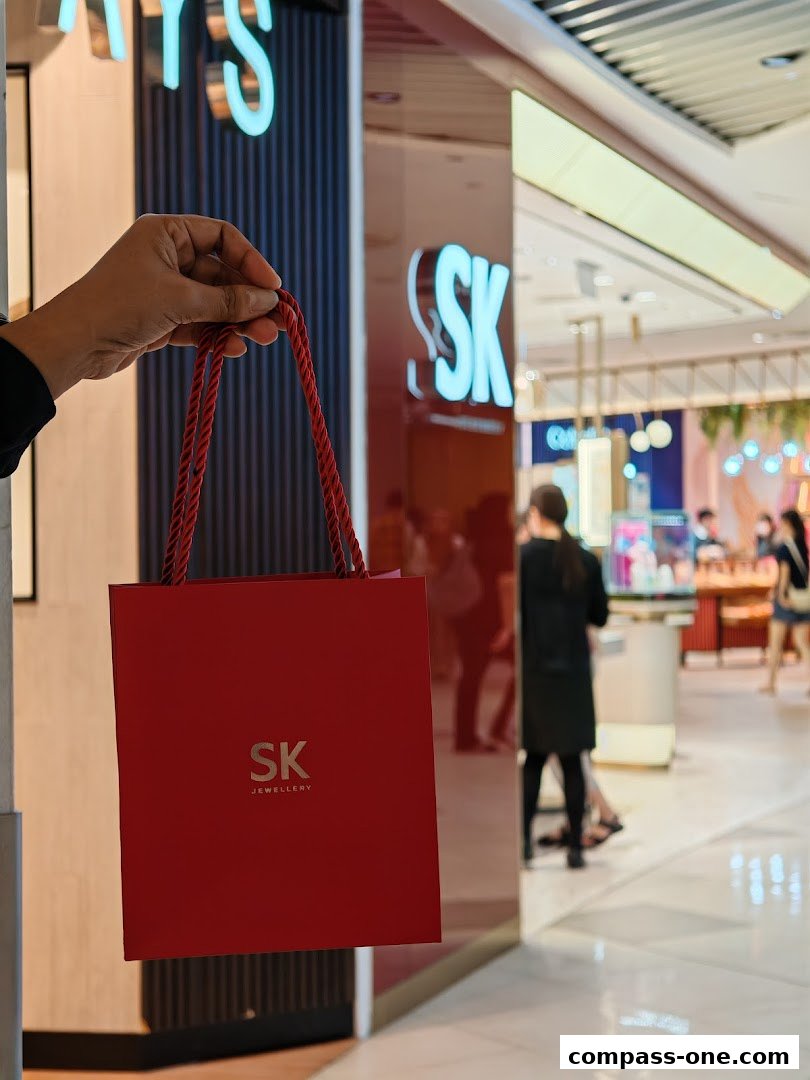 A hand holds a red SK Jewellery shopping bag in front of the store.