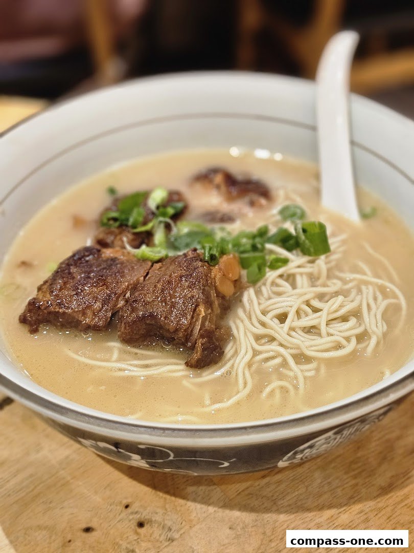 A close-up shot of a steaming bowl of rich, creamy noodle soup with tender braised beef and chopped scallions.