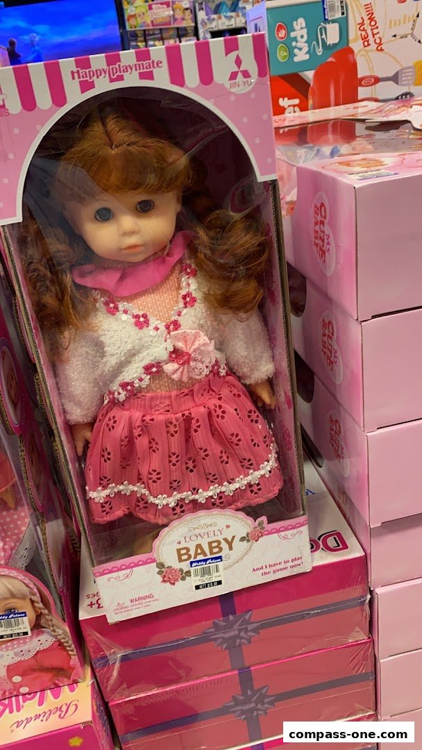 A doll in a pink dress and white sweater is displayed in a box, with other toy boxes stacked behind it.