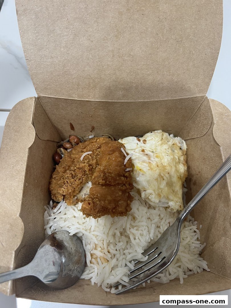 A takeaway box filled with Nasi Lemak, featuring rice, fried chicken, egg, and anchovies.